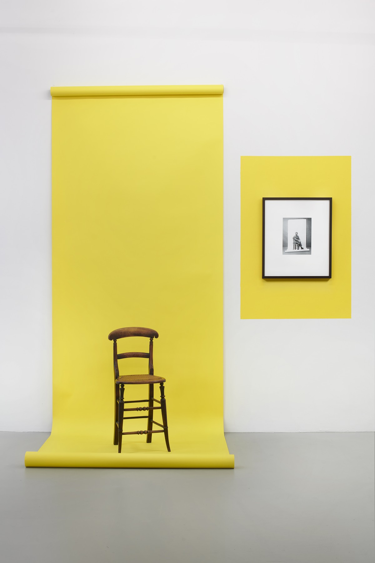 Barbara Bloom. Homage to JL Godard (Corrected). 1986-2021. Seamless yellow backdrop paper. Framed facsimile photograph of J-L Godard by F. C. Gundlach from 1961. Victorian wooden and cane child’s post